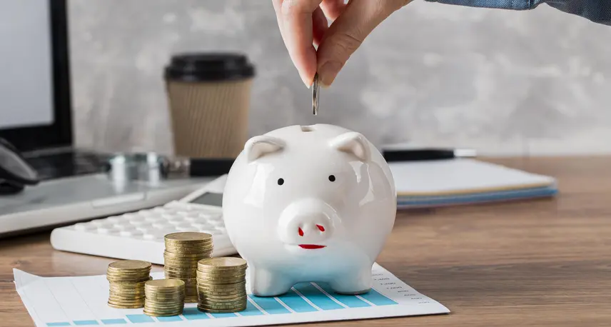 A piggy bank with some coins, a calculator and a coffe cup in the background